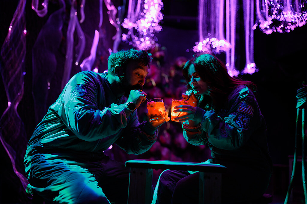 Guests enjoying cocktails in night-time scene inside Avora with glowing plants and immersive lighting.